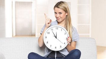 Eine Frau sitzt auf einem Sofa und hält eine Wanduhr mit dem Zeiger auf 3 Uhr. Sie zeigt mit dem Finger nach oben und lächelt.