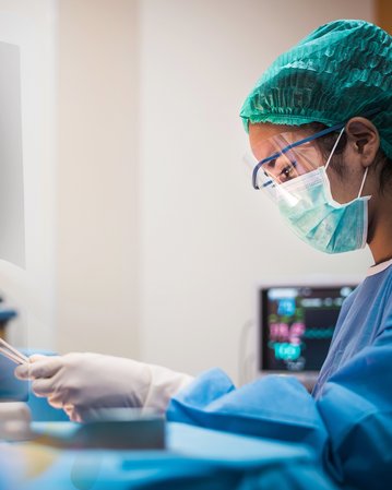 Eine medizinische Fachkraft in Schutzkleidung überwacht Vitalzeichen auf einem Monitor. Der Bildschirm zeigt Herzfrequenz, Blutdruck und Sauerstoffsättigung an. Die Person hält ein Instrument in der Hand und konzentriert sich auf die Daten.