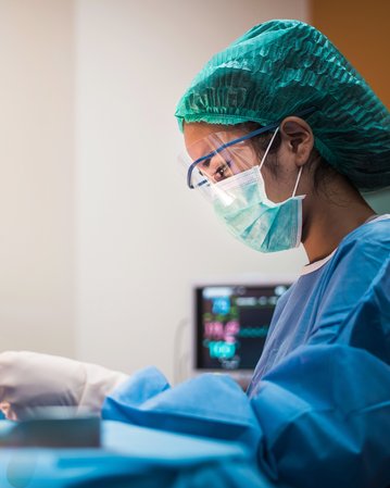 Eine medizinische Fachkraft in Schutzkleidung überwacht Vitalzeichen auf einem Monitor. Der Bildschirm zeigt Herzfrequenz, Blutdruck und Sauerstoffsättigung an. Die Person hält ein Instrument in der Hand und konzentriert sich auf die Daten.