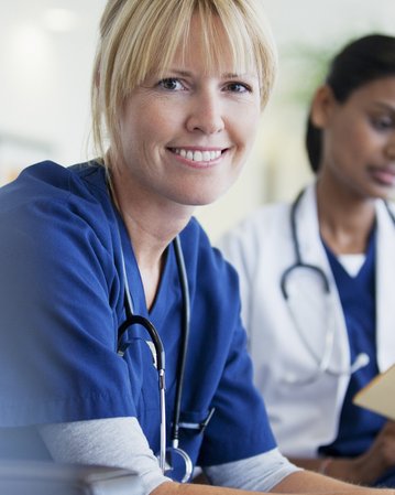 Eine lächelnde Krankenschwester in blauer Uniform mit einem Stethoskop um den Hals, während eine andere Person im Hintergrund arbeitet.