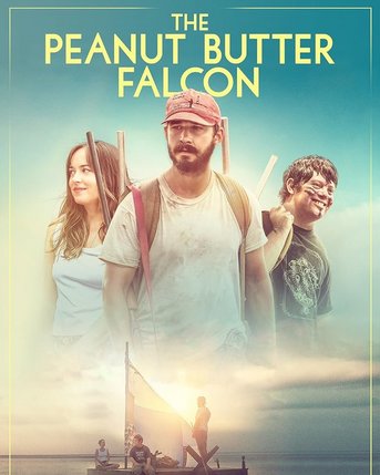 Drei Hauptfiguren aus dem Film "The Peanut Butter Falcon" stehen vor einem hellen Himmel, während ein Boot im Wasser zu sehen ist.