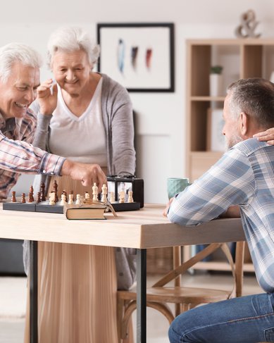 Vier ältere Menschen spielen Schach an einem Tisch. Zwei Personen beobachten das Spiel aufmerksam, während eine Person einen Zug macht. Ein weiteres Mitglied der Gruppe sitzt entspannt und schaut zu.
